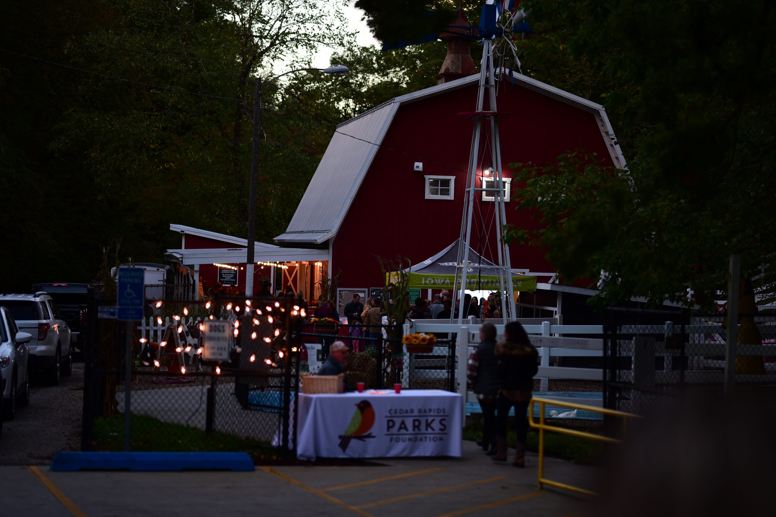 This is a picture of the 2022 Beers in the Barnyard event. The Old McDonald Farm is in the background along with an Iowa Brewing Co. Tent. In the foreground is a table adorned with a Cedar Rapids Parks Foundation tablecloth. There are lights on the fencing, and people are scattered around the event.